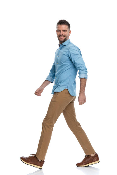 sexy hombre de mediana edad con zapatillas de deporte en camisa de mezclilla con pantalones chinos pisando y sonriendo sobre fondo blanco - Foto, imagen