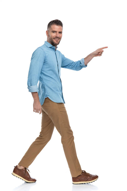barbudo chico de mediana edad sonriendo y señalando el dedo mientras camina aislado en una pose vista lateral sobre fondo blanco - Foto, imagen