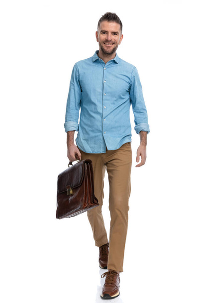 uomo di mezza età non rasato con camicia di jeans che tiene la valigia e sorride su sfondo bianco mentre cammina isolato in studio - Foto, immagini