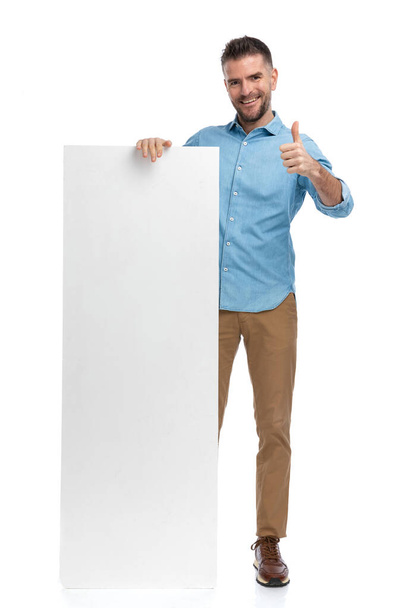 guapo hombre sosteniendo la cartelera y haciendo pulgares hacia arriba gesto mientras sonríe sobre fondo blanco - Foto, imagen