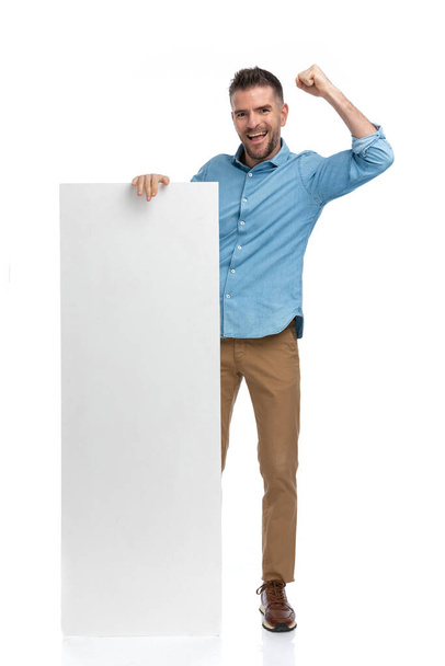 full body picture of young excited man with arm in the air holding board and celebrating success on white background - Fotó, kép