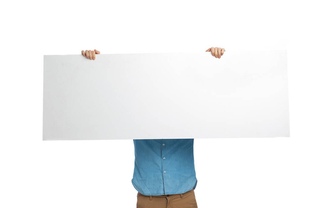 man in jeans shirt holding in the air and covering face with long empty board on white background in studio - Photo, Image