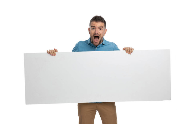 σοκαρισμένος casual τύπος με μεγάλη λευκή πινακίδα στα χέρια είναι έκπληκτος από τη διαφήμιση σε λευκό φόντο στο στούντιο - Φωτογραφία, εικόνα