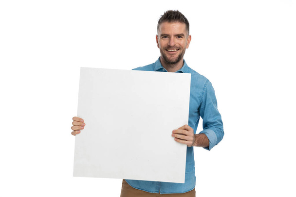 stolzer unrasierter Mann, der eine Tafel hält und Werbung zeigt, während er lächelt und in seiner Arbeit auf weißem Hintergrund zuversichtlich ist - Foto, Bild