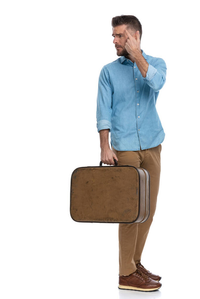 расстроен молодой человек с багажом указывая пальцем в висок, стоя в очереди и злясь, глядя назад через плечо на белом фоне - Фото, изображение