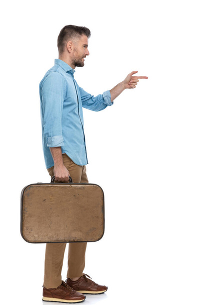 concept of casual young man with luggage enjoying holiday, going to vacation, waiting in line to buy tickets for flight and pointing finger on white background - Foto, Imagen