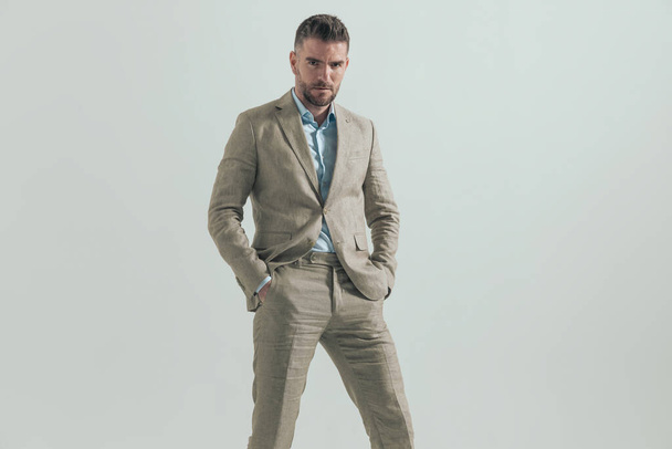 retrato de hombre de negocios barbudo guapo cogido de la mano en bolsillos y posando delante de fondo gris  - Foto, Imagen