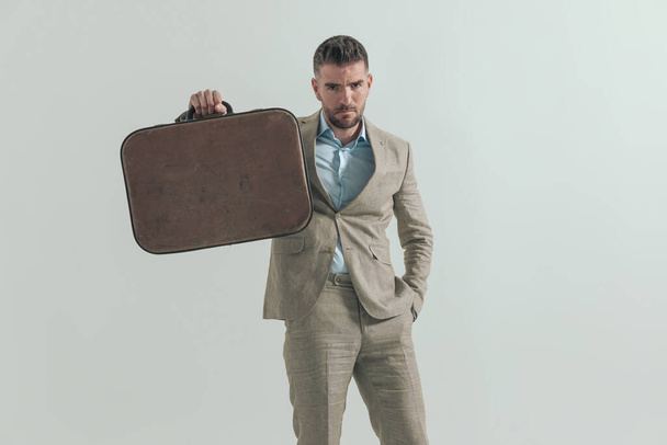 middle aged man with hand in pocket holding and showing luggage asking for some days off to go on vacation, posing in front of grey background - Photo, Image
