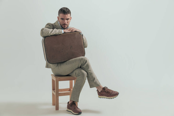 αξύριστος νεαρός άνδρας με κοστούμι και αποσκευές κάθεται σε ξύλινη καρέκλα και φεύγει από την πόλη στις ημέρες του μακριά από την εργασία - Φωτογραφία, εικόνα