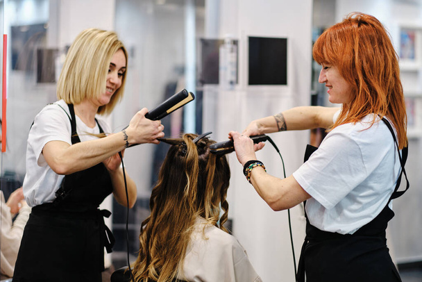 Dwóch profesjonalnych fryzjerów korzystających ze wspólnej pracy podczas stylizacji włosów klienta w salonie piękności. - Zdjęcie, obraz