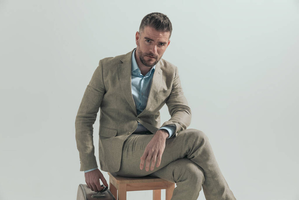 hombre de traje con equipaje sentado en una silla de madera y tomando algunos días libres para ir de vacaciones, posando sobre fondo gris - Foto, Imagen