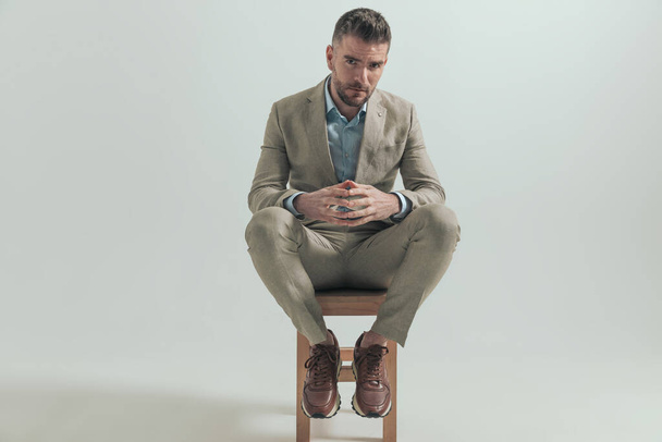 sexy junger Geschäftsmann mit Ellbogen auf Knien, die Finger berühren und auf einem Holzstuhl auf grauem Hintergrund im Studio sitzen - Foto, Bild