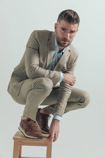 hombre de negocios sin afeitar agachado en una silla de madera, sosteniendo el codo en la rodilla y posando sobre un fondo gris en el estudio - Foto, Imagen