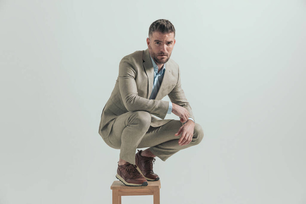 attractive man in his forties holding elbows on knees and crouching on wooden chair in front of grey background in studio - Photo, image