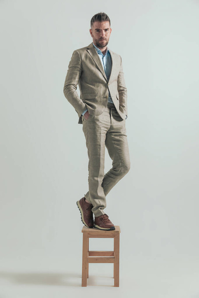 бізнесмен середнього віку в костюмі на сірому фоні, що стоїть з руками в кишенях на дерев'яному стільці в студії
 - Фото, зображення
