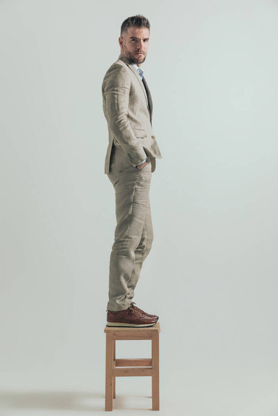 πλαϊνή άποψη του νεαρού επιχειρηματία με γενειάδα στέκεται σε ξύλινη καρέκλα με τα χέρια στις τσέπες και σκέφτεται να βγει από τη ζώνη άνεσής του - Φωτογραφία, εικόνα