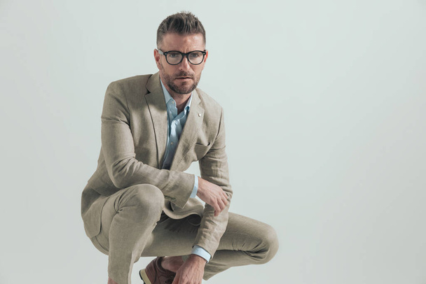 sexy hombre de negocios en traje con camisa de cuello abierto y gafas agachándose y sosteniendo el codo en la rodilla en una pose de moda sobre fondo gris - Foto, imagen