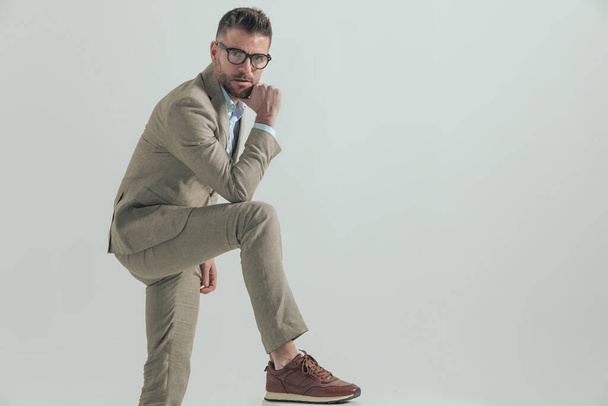 boční pohled na chytrý podnikatel s brýlemi drží ruku na bradě a myšlení, zatímco drží loket na koleni a nohu nahoru na šedém pozadí - Fotografie, Obrázek