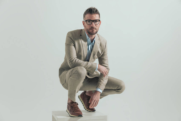 handsome unshaved man in his 40s with elbow on knee crouching and squatting while looking up dressed in a brown suit with open collar shirt - Photo, Image