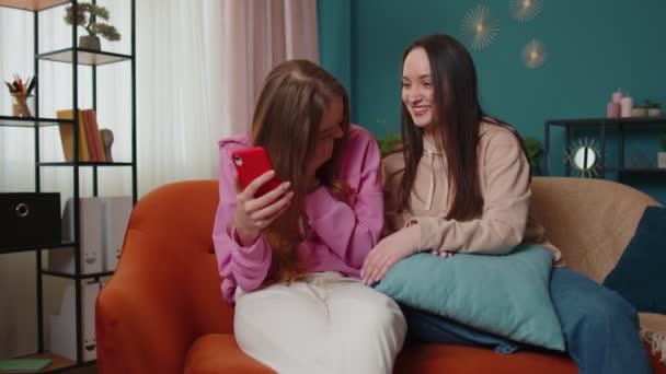 陽気な女の子の友人の兄弟は自宅でソファの上でスマートフォンで写真を見て、ソーシャルメディアを楽しむ - 映像、動画