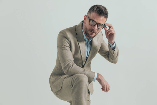 serious man with beard holding elbow on knee fixing glasses and posing in front of grey background - Photo, Image