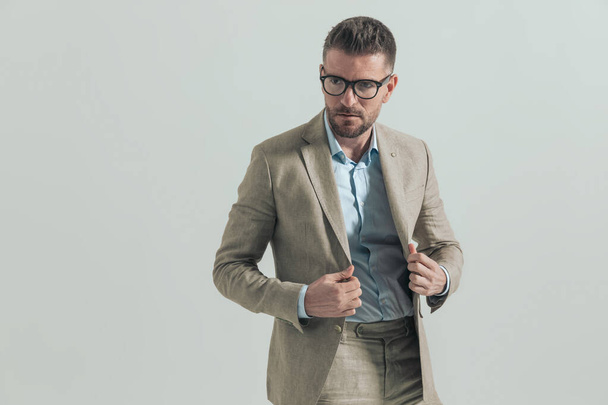 πορτρέτο του αξύριστου μεσήλικα επιχειρηματία με γυαλιά σε γκρι φόντο τακτοποίηση κοστούμι και κοιτάζοντας μακριά  - Φωτογραφία, εικόνα