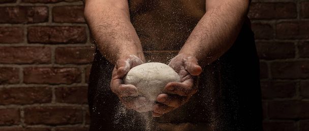 Beautiful and strong men's hands knead the dough from which they will then make bread, pasta or pizza. A cloud of flour flies around like dust. Food concept - Foto, imagen