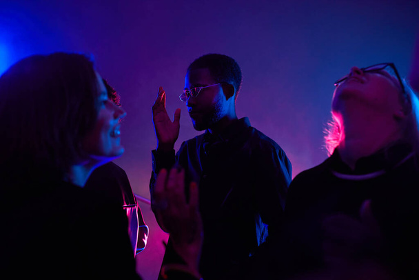 Πλάτη φωτισμένο πορτρέτο του μαύρου νεαρού άνδρα που χορεύει στο πλήθος ενώ διασκεδάζει σε smoky club φωτισμένο από νέον, αντιγραφή χώρου - Φωτογραφία, εικόνα