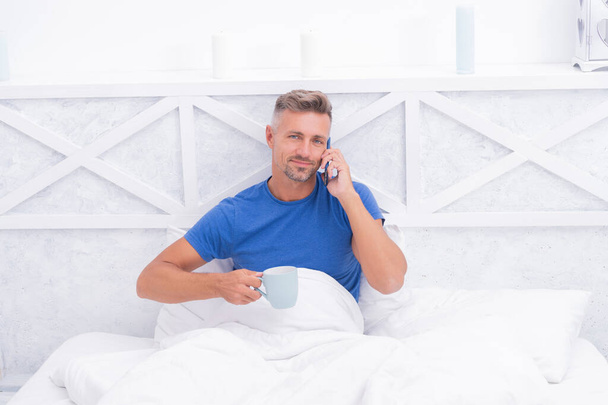 Happy man talking on mobile phone while drinking morning tea or coffee in bed, call - Fotografie, Obrázek
