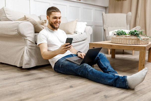 young man at home on the floor near the sofa working on a laptop with a phone. - Foto, Bild