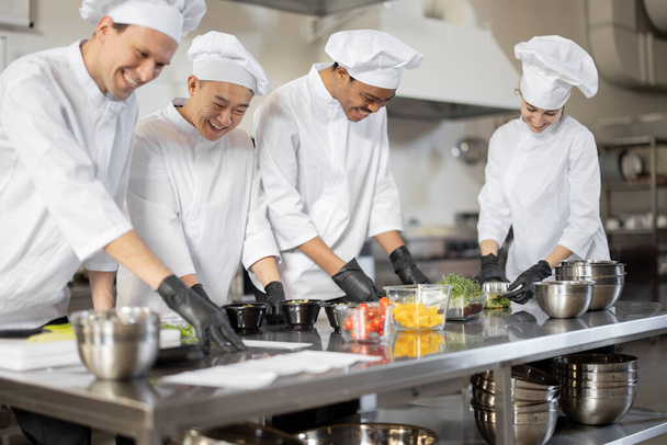 Equipo multirracial de cocineros mezclando ingredientes para llevar comida en cocina profesional - Foto, imagen