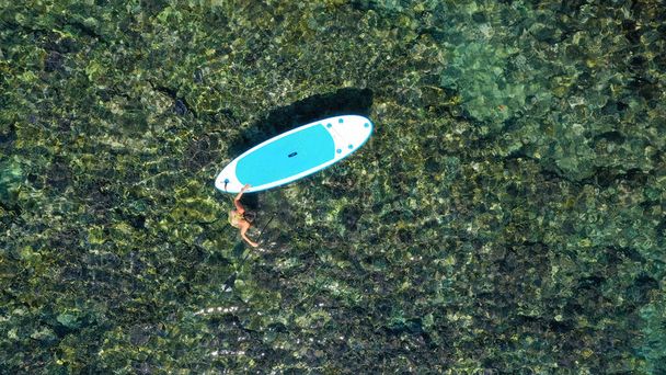Z góry na dół, lecące nad młodą kobietą w zielonym bikini w pobliżu paddleboard. Kobieta w turkusowym morzu ze stojącym wiosłem. Nadmuchiwana deska SUP na krystalicznie czystej wodzie oceanu. Widoczne dno skaliste - Zdjęcie, obraz