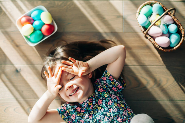 Happy smiling little girl lying on a floor with pastel colored Easter eggs in a basket - Photo, Image