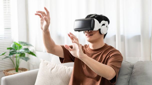 Technology Concept El hombre con su camiseta marrón y vaqueros azul claro con un casco de realidad virtual sentado en el sofá. - Foto, Imagen