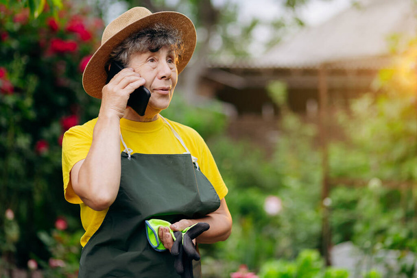 Femme jardinière senior dans un chapeau travaillant dans sa cour et parle au téléphone. Le concept de jardinage, de culture et d'entretien des fleurs et des plantes. - Photo, image