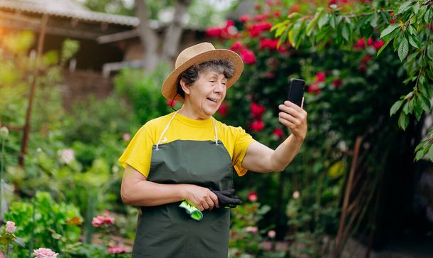 Oudere tuinier in een hoed die in haar tuin werkt en aan de telefoon spreekt. Het concept van tuinieren, groeien en verzorgen van bloemen en planten. - Foto, afbeelding