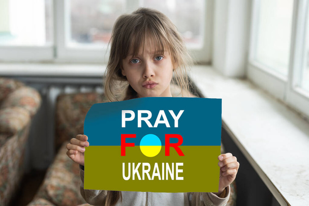 Sad little girl covered with Ukraine flag. Concept of standing with Ukrainian nation in war with Russia - Photo, Image