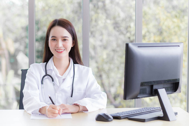 Professional Asian woman doctor wears medical coat and stethoscope while she is writing something on the paper and working in office room which she looking at the camera at hospital with health check concept. - Foto, Imagem