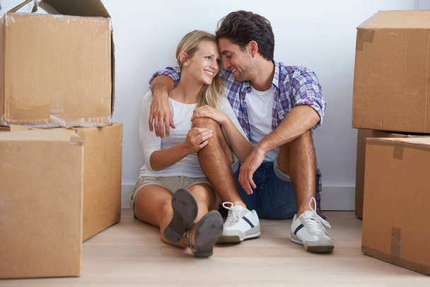 Yeni aşk yuvalarında mutlular. Mutlu genç bir çift yeni evlerinde yerde oturuyor ve kutularla çevrelenmiş bir şekilde birbirlerine sarılıyorlar.. - Fotoğraf, Görsel