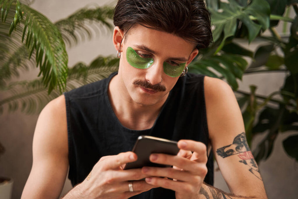 Attentive stylish man with under eye patches sitting at home and surfing net while relaxing - Фото, зображення