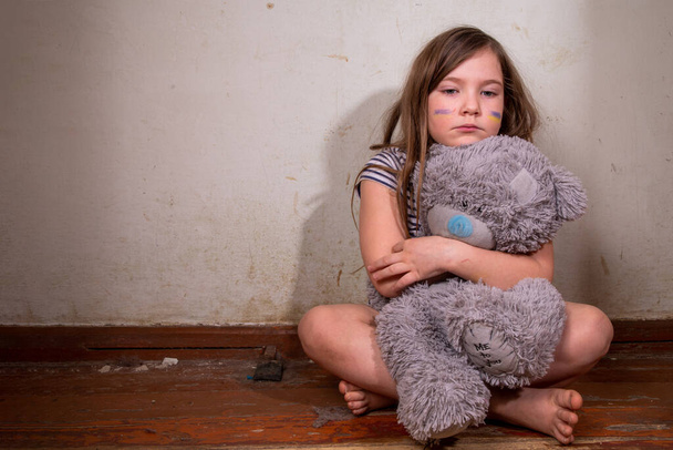 Stop war in Ukraine concept. Little Ukrainian blond girl, ukrainian flags painted on cheeks, with teddy bear toys in a ruined old house. Save the children, close the sky of Ukraine, humanitarian aid disaster concept - Photo, Image