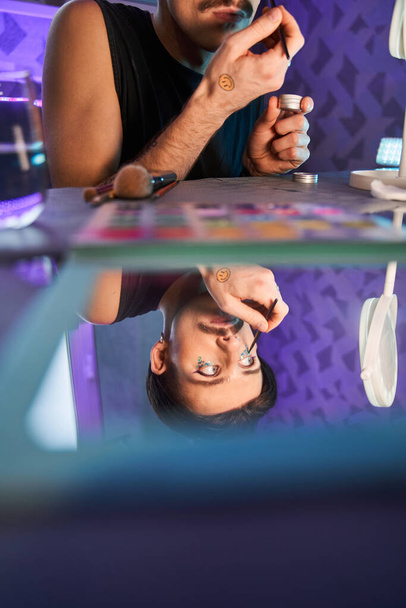 Metrosexual man applying make-up at his eyes while sitting at the table at home - 写真・画像