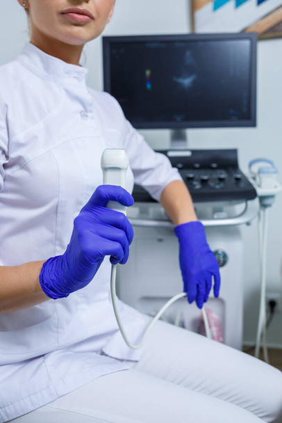 Genç bir kadın stajyer doktor, elinde ultrason cihazı tutan beyaz bir takım elbise ve tıbbi eldiven giyiyor. Ultrason kontrolleri. seçici odak - Fotoğraf, Görsel
