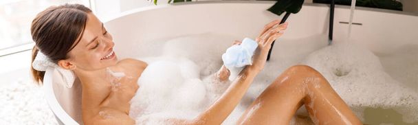 Прекрасная молодая женщина купается, используя губку и заботясь о своей коже, расслабляясь в ванной - Фото, изображение