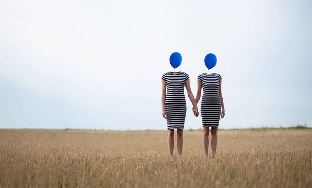 surrealistinen kuva kahdesta naisesta, jotka seisovat käsi kädessä kentällä, heidän päänsä korvataan sinisillä ilmapalloilla, kopioi tilaa - Valokuva, kuva