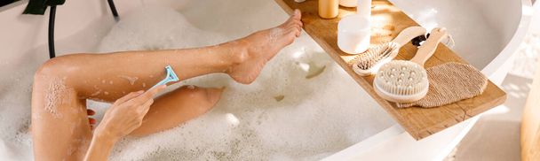 Legs of young woman shaving with disposable shaving razor while bathing. Body care cosmetics on wooden shelf over bathtub - Photo, Image