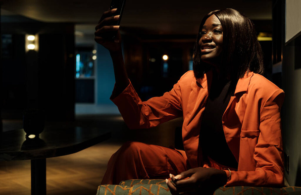 Portrait of young black woman using smartphone for selfportrait in a lobby hotel. She is wearing an orange jacket. - Fotografie, Obrázek