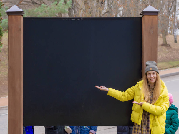 Улыбающаяся девушка в желтой куртке указывает на пустой стенд в осеннем городском парке. - Фото, изображение