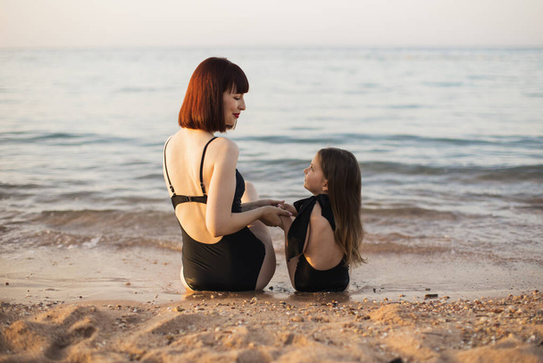 Attractive woman mother with red hair and her little cute daughter sitting on the sea beach. - Valokuva, kuva