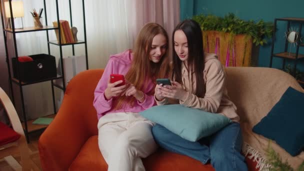Veselé dívky přátelé sourozenci sledování fotografií na smartphone na pohovce doma, těšit sociální média - Záběry, video